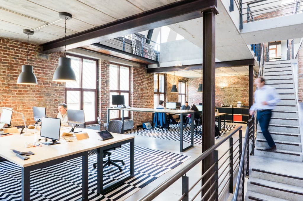 An open office with brick walls and long desks, a colleague heading up the stairs
